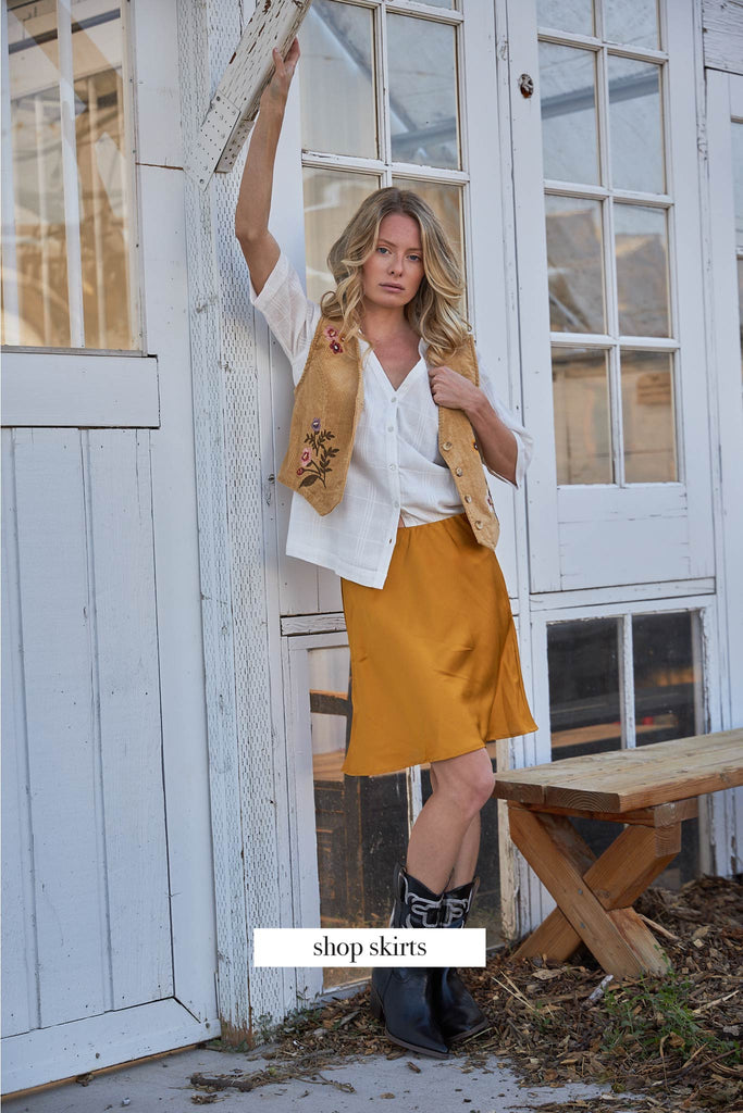 Text over image states "Shop Skirts". A women dressed in white shirt, yellow vest, and gold skirt.